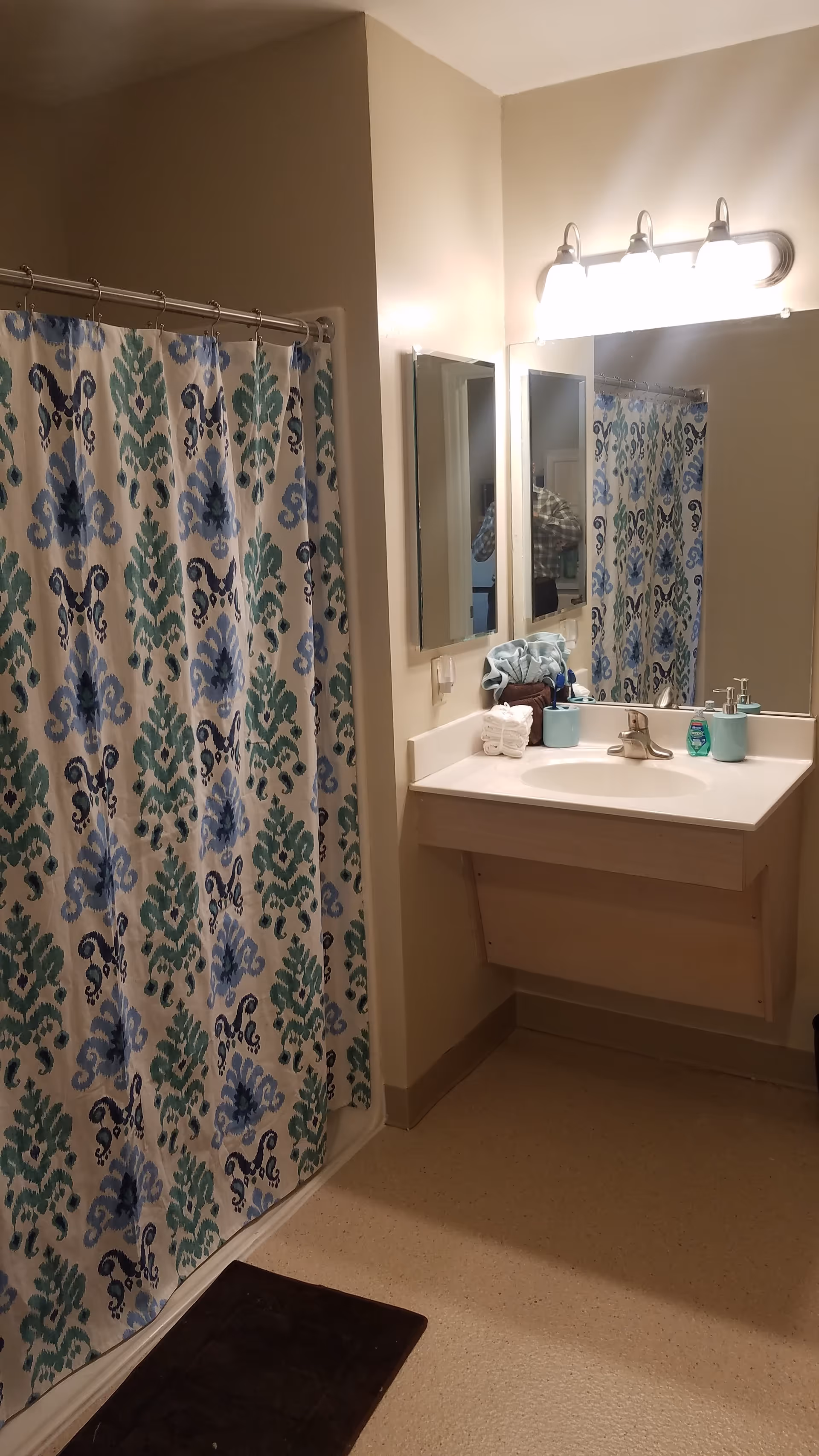 Bathroom with a patterned shower curtain in shades of blue and green, a white sink with a silver faucet, a large mirror above the sink, and a light fixture with three bulbs. There are towels and toiletries on the sink counter.