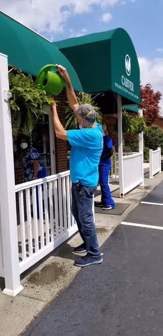A man wearing a blue shirt and jeans waters hanging plants outside the entrance of Carver Rehabilitation & Living Center. Two other people, one in blue scrubs and another partially visible inside the building, are nearby. The building has a green awning with the facility's name and logo. The scene is outdoors on a sunny day.