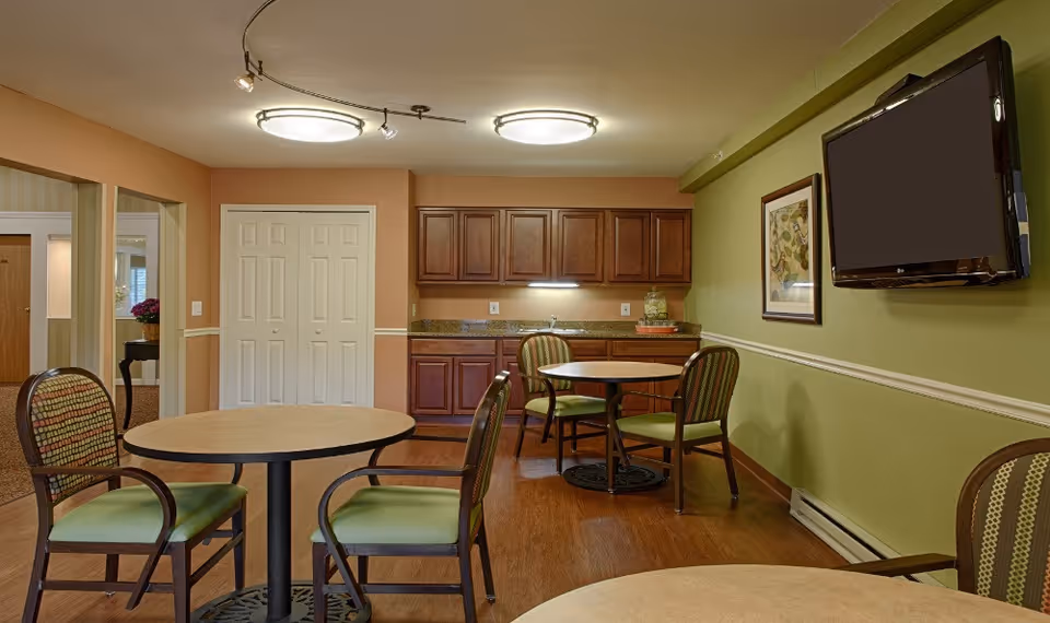 Interior view of a common area with two round tables and several chairs with green cushions. The room has wooden flooring, a small kitchenette with wooden cabinets and a sink, a wall-mounted flat screen TV, and a framed picture on the green wall.