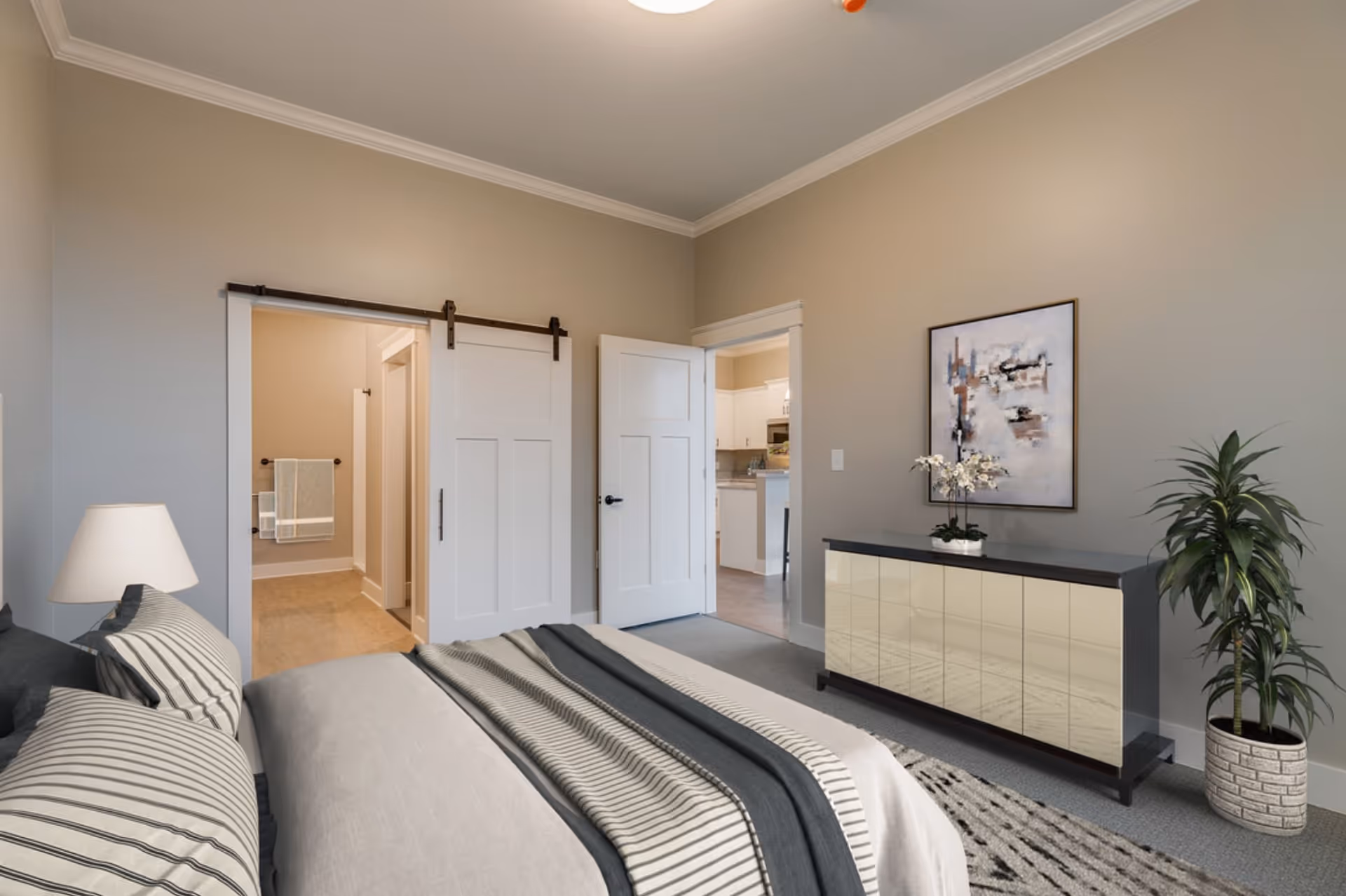 A bedroom with a neatly made bed featuring striped pillows and blankets. There is a bedside lamp on the left side. The room has beige walls and a gray carpet. A modern dresser with a reflective front is against the right wall, decorated with a small plant and a framed abstract painting above it. A potted plant is placed on the floor next to the dresser. Two doors are visible: one sliding barn door leading to a bathroom with towels hanging on a rack, and another door opening into a kitchen area.