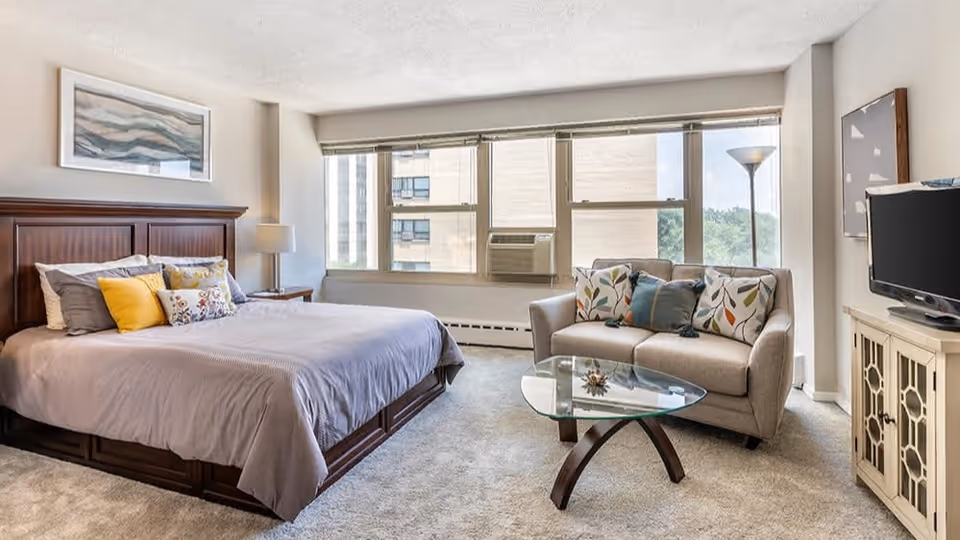 A bright bedroom with a large bed featuring multiple pillows, a wooden headboard, a nightstand with a lamp, a beige loveseat with decorative pillows, a glass coffee table, a floor lamp, a TV on a cabinet, and large windows letting in natural light.