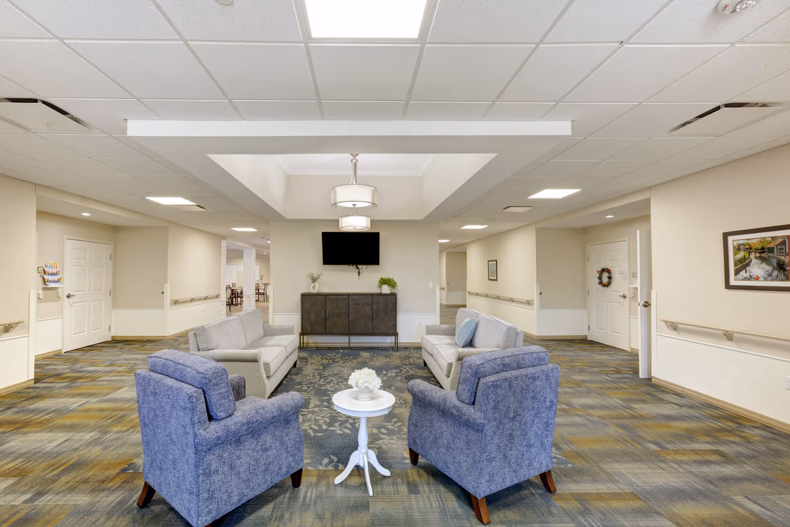 A bright and spacious common area in a senior living facility featuring two gray sofas and two blue armchairs arranged around a small white round table with a flower centerpiece. The room has a patterned carpet, neutral-colored walls, a ceiling with recessed lighting and a central hanging light fixture. A flat-screen TV is mounted on the wall above a dark cabinet, and there are doors and hallways leading to other parts of the facility.