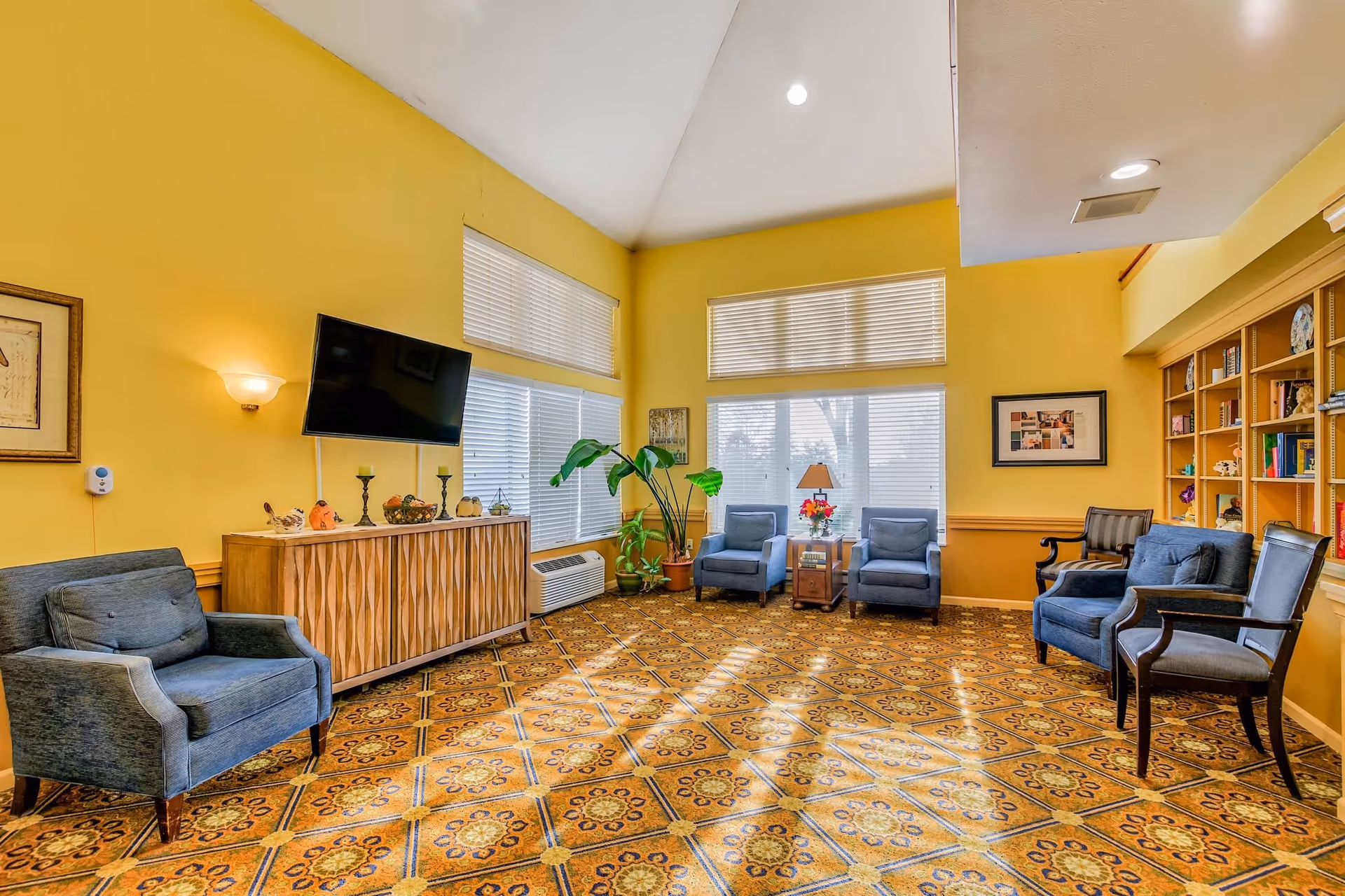 A bright living room with yellow walls and a patterned carpet. The room features several blue upholstered chairs arranged around the space, a wooden cabinet with decorative items on top, a wall-mounted flat-screen TV, large windows with blinds, a potted plant, a table with a lamp and flowers, and built-in bookshelves filled with books and decorative pieces.