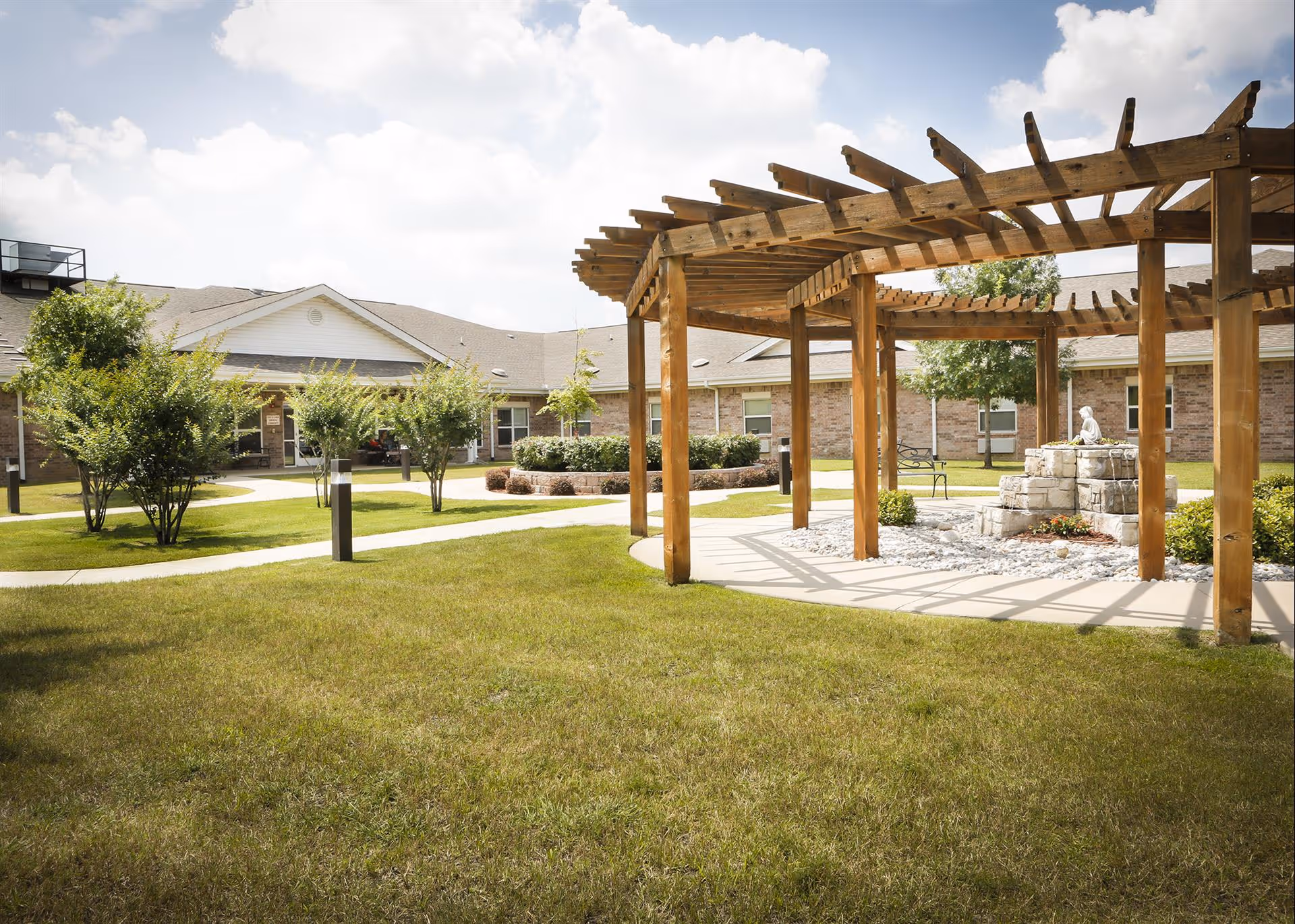 Outdoor courtyard area at Bethesda Gardens Fort Worth featuring a wooden pergola, a stone water fountain with a small statue, green grass, trees, shrubs, and a brick building in the background under a partly cloudy sky.
