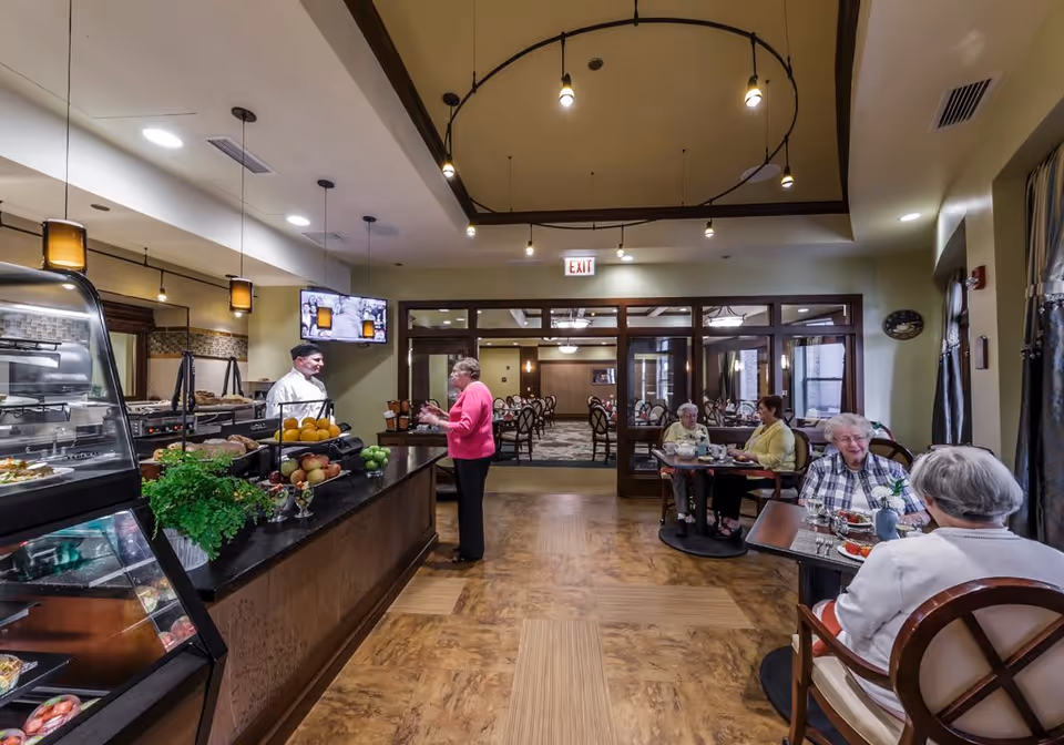 Residents sit at tables in a senior living dining room while a staff member talks with a woman at a serving counter.