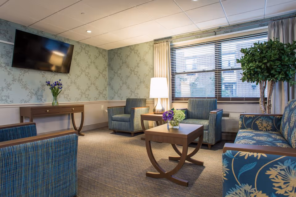 A cozy living room area in a senior living facility with patterned wallpaper, a wall-mounted flat-screen TV, a wooden console table with a vase of flowers, two blue upholstered armchairs, a wooden coffee table with a small flower arrangement, a floor lamp, a large window with blinds and curtains, and a patterned sofa next to a tall indoor plant.
