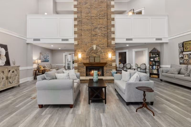 A spacious living room with two gray sofas facing each other across a dark wooden coffee table. A brick fireplace with a fire burning is centered on the far wall, flanked by two wall sconces. The room has light gray walls, wood flooring, and white trim. There is a wooden side table next to one sofa, a bookshelf with books and framed photos, and additional seating in the background.
