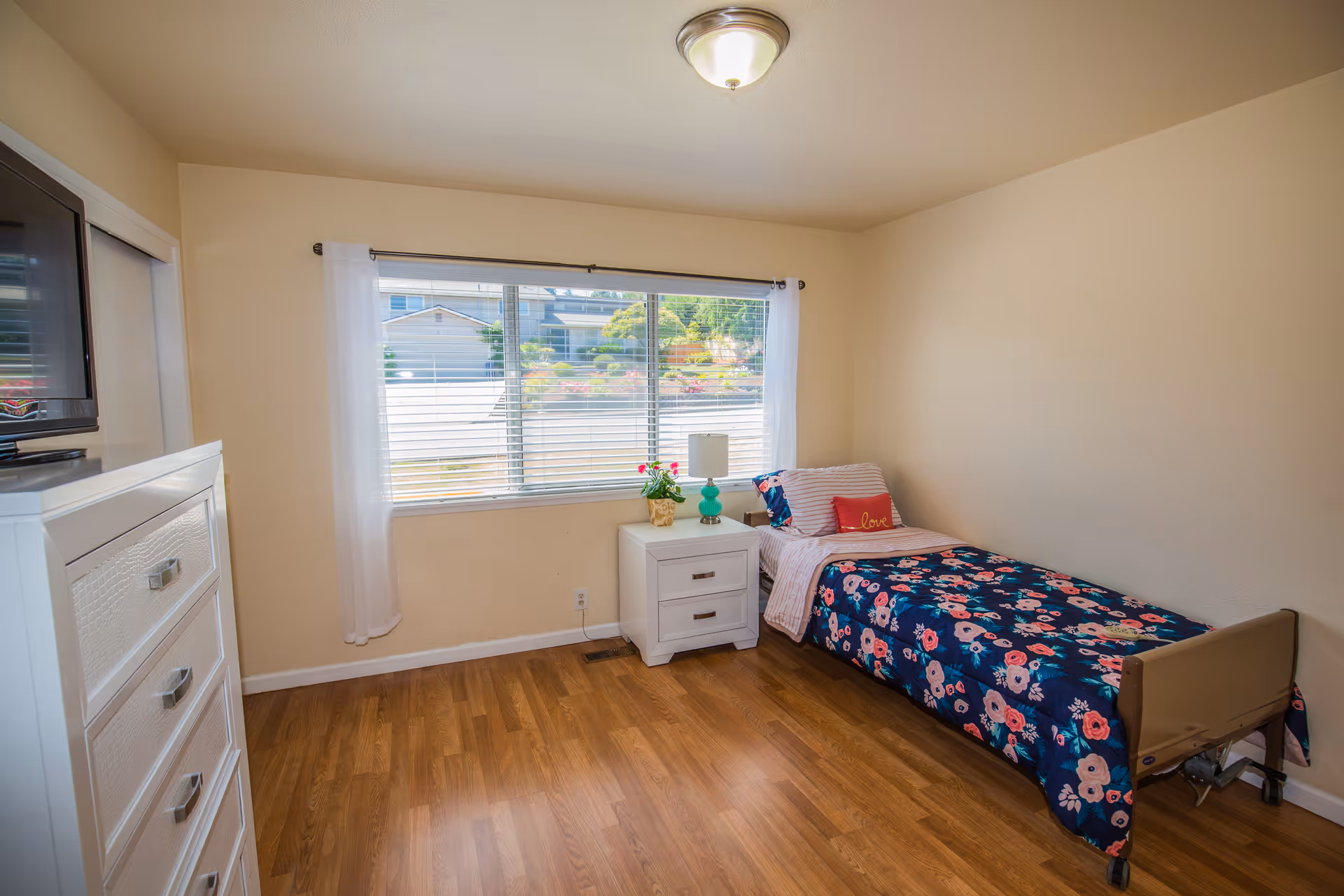 A small bedroom with a single bed covered in a dark blue floral bedspread and a red pillow with the word 'love'. Next to the bed is a white nightstand with a turquoise lamp and a small plant. A large window with white curtains lets in natural light. There is a white dresser with a TV on top on the left side of the room. The floor is wooden and the walls are painted beige.
