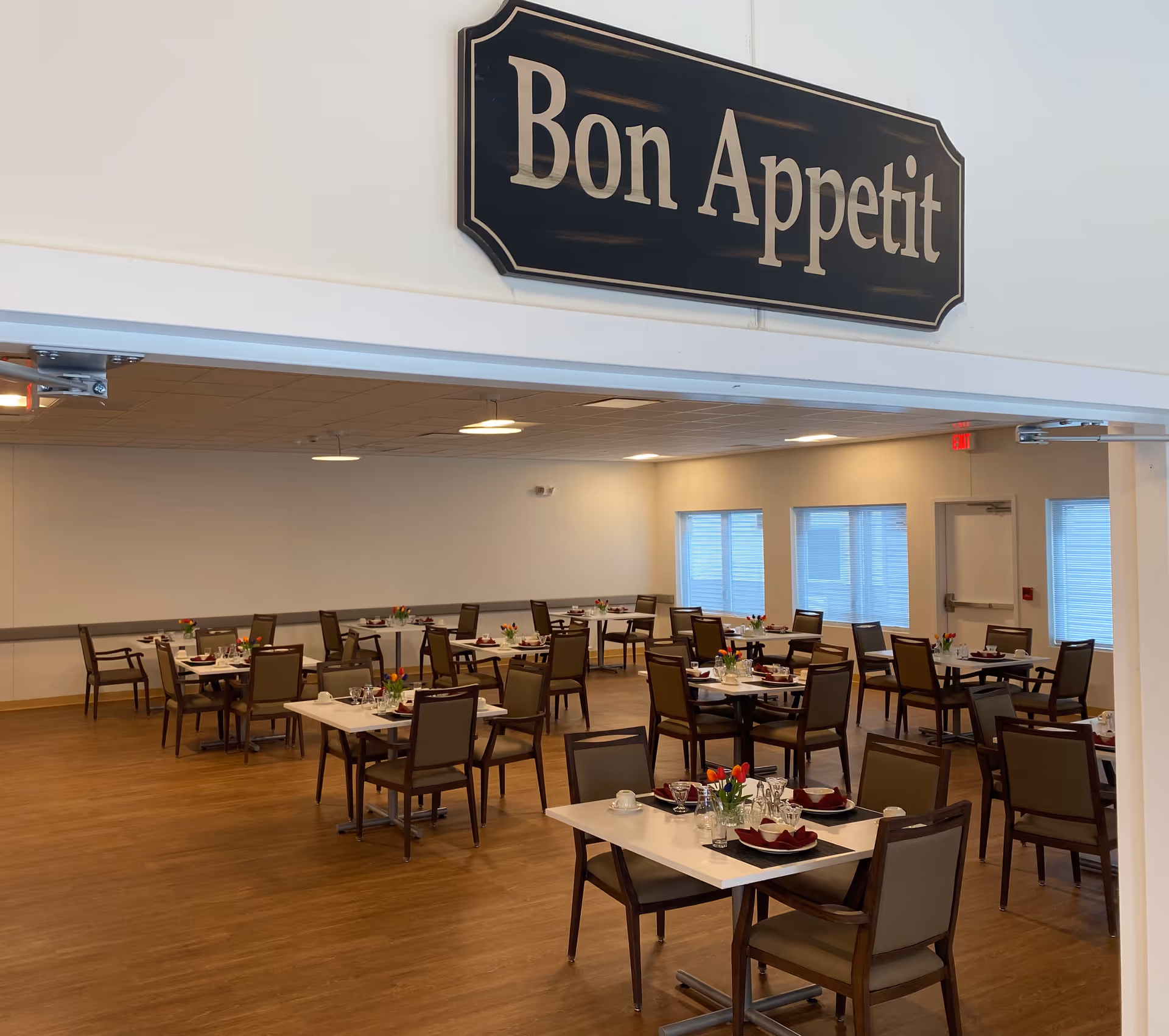 A dining room with multiple tables set with plates, glasses, napkins, and small flower arrangements. The room has wooden flooring, beige walls, and several windows letting in natural light. A sign above the entrance reads 'Bon Appetit'.