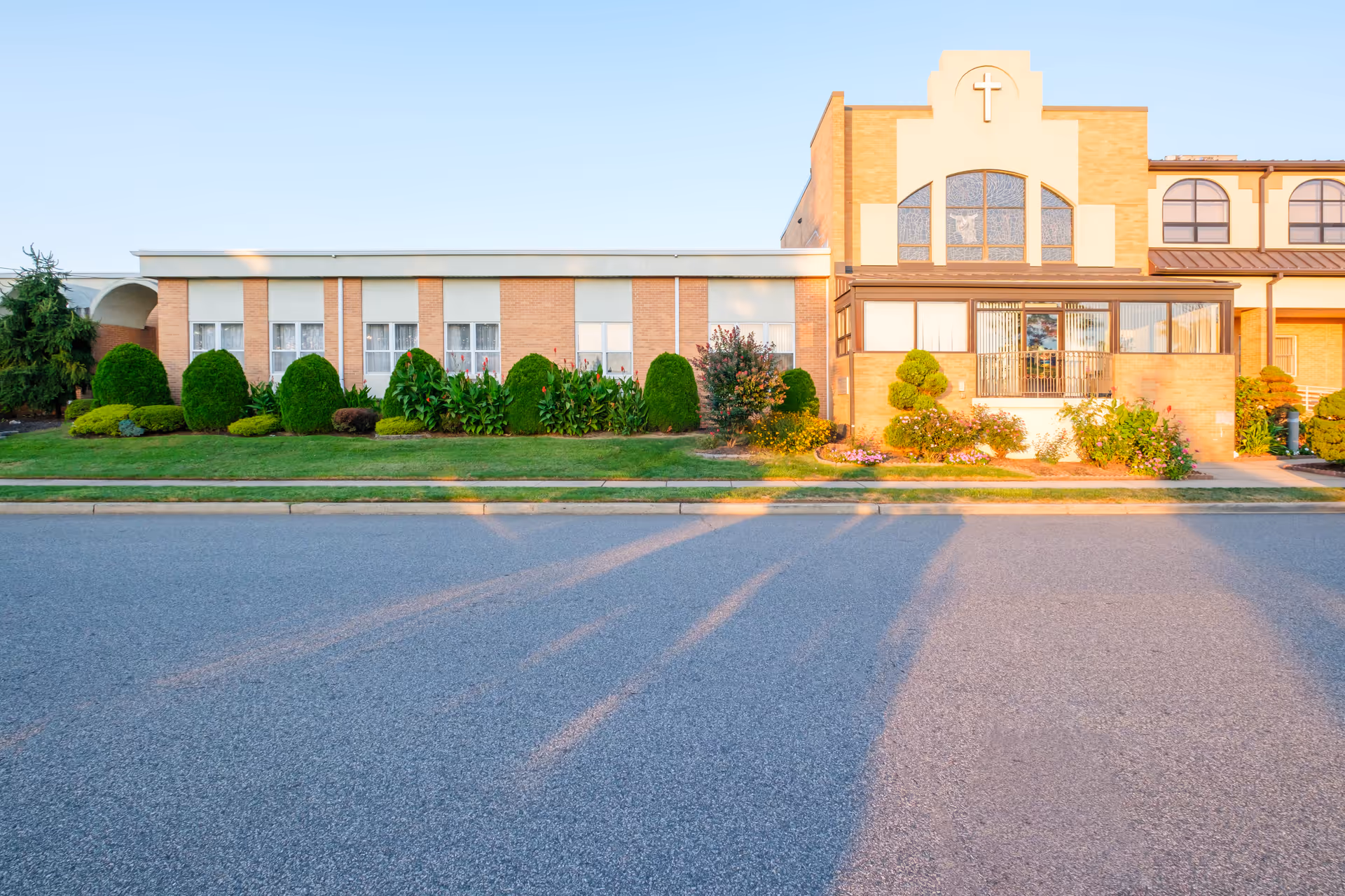 Exterior view of St Joseph's Senior Home - Assisted Living and Nursing Center building with a well-maintained lawn, shrubs, and flowers in front. The building features brick and light-colored walls with a cross symbol above a large window.