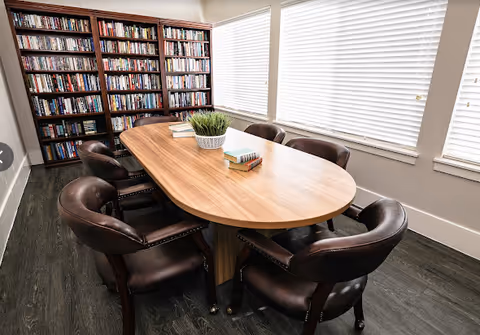 Oval wooden table surrounded by six leather chairs in a bright room with bookshelves and window blinds.