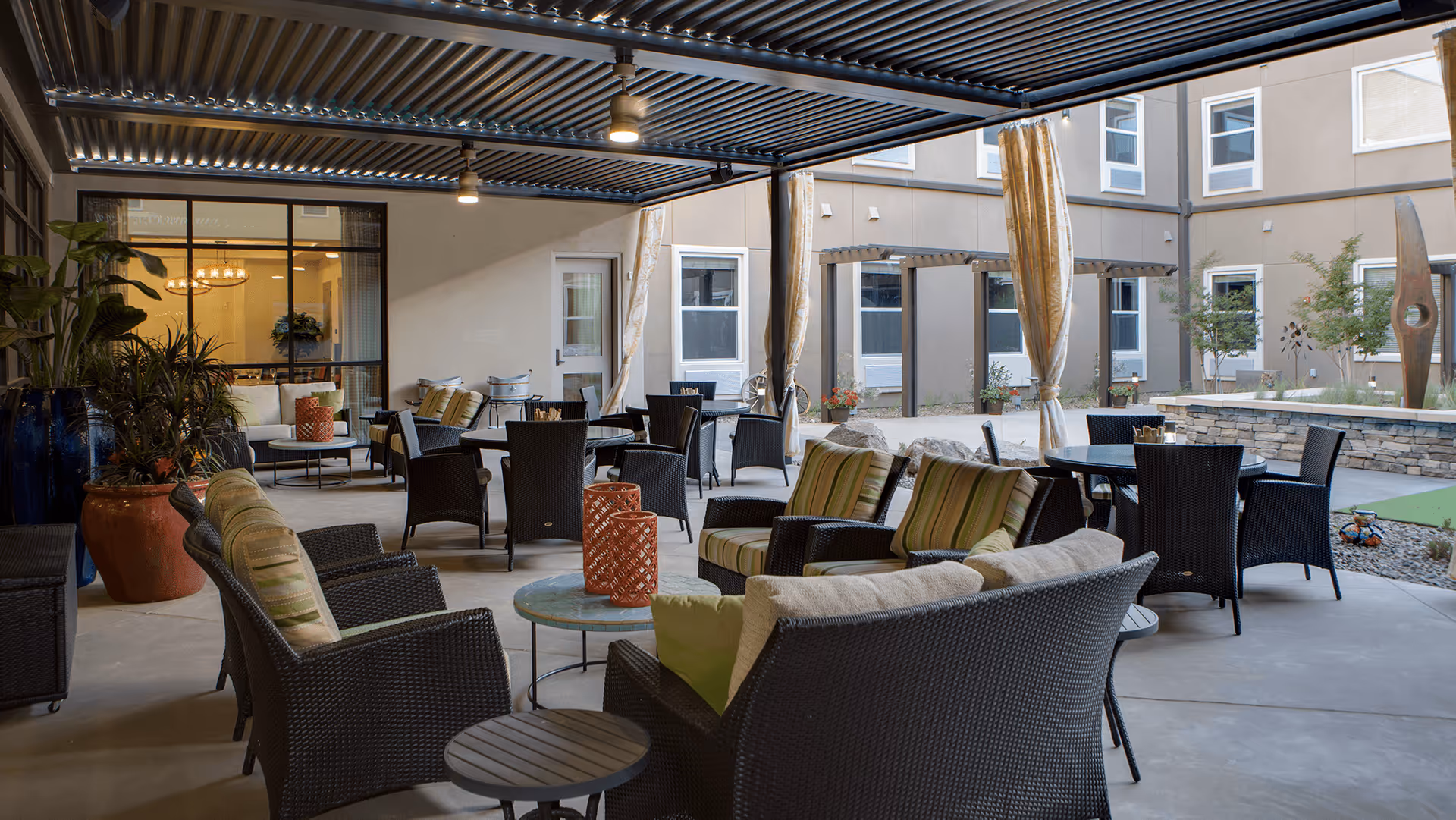 Covered outdoor patio area with multiple wicker chairs and sofas with cushions arranged around small tables. Large potted plants are placed on the left side, and the patio overlooks a courtyard with windows and some greenery. The area is well-lit with ceiling lights and has a modern, comfortable seating arrangement.