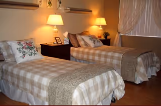 A cozy bedroom with two single beds covered in beige and white checkered bedspreads, each with decorative pillows. Between the beds is a dark wooden nightstand with two lit table lamps, framed photos, and a small plant. The room has warm lighting and a window with sheer curtains.