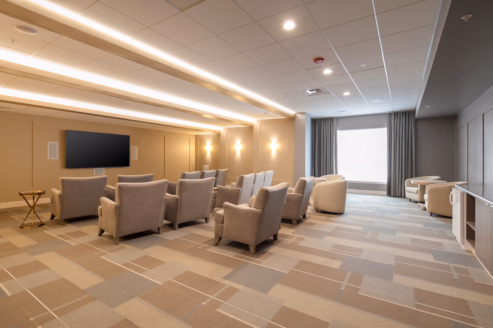 A modern lounge area with multiple beige armchairs arranged in rows facing a large flat-screen TV mounted on a beige wall. The room has soft lighting with wall sconces and recessed ceiling lights. There is a large window with curtains on the right side, and the floor is covered with a patterned carpet in neutral tones.