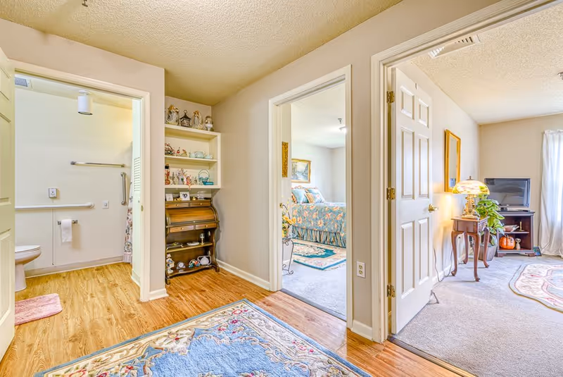 Bright interior hallway of a senior living apartment showing an open bathroom, a bedroom, and a living area with furniture and decorative rugs.