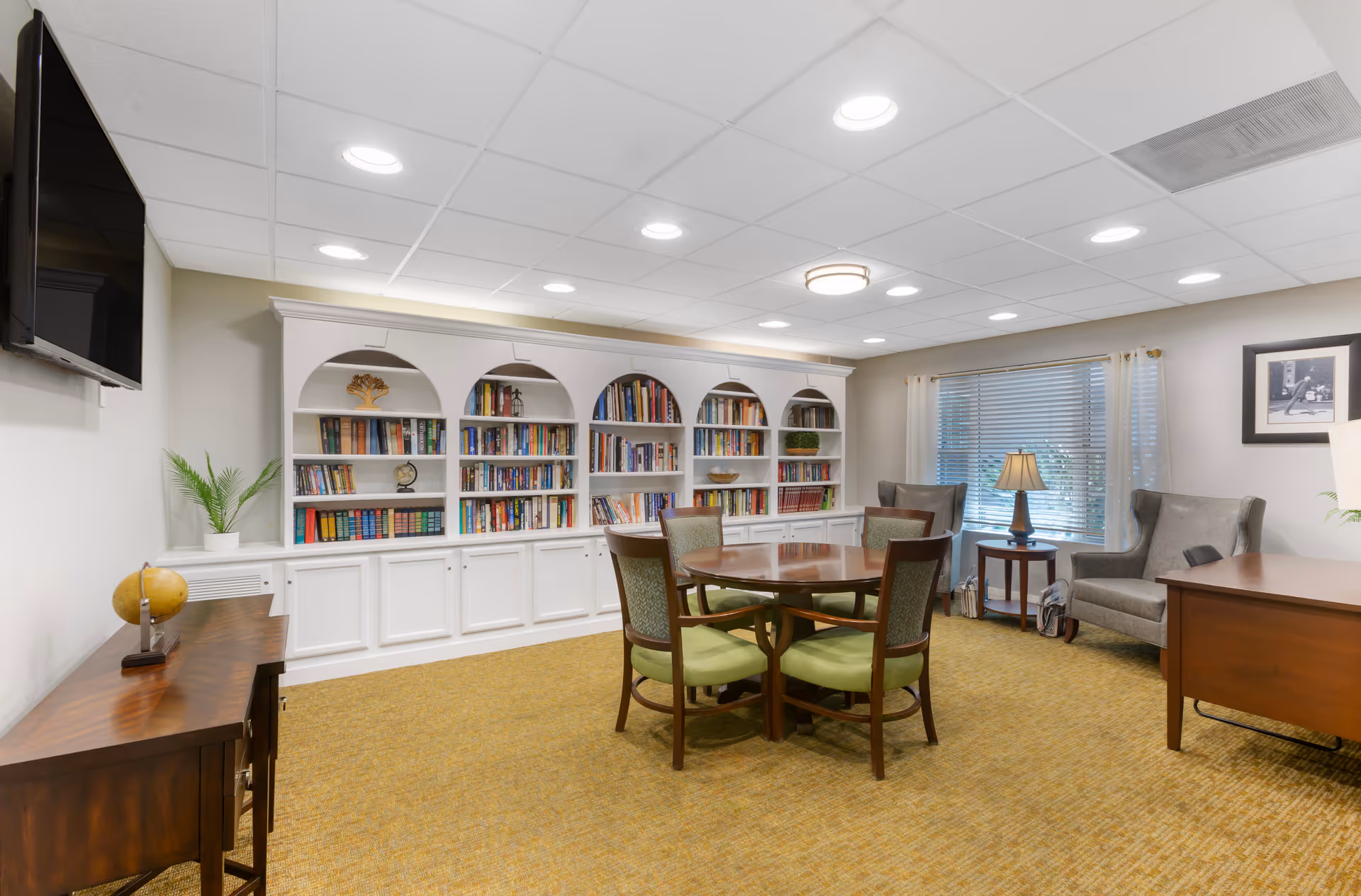 Well-lit common room with built-in white bookshelves, a round table with green-upholstered chairs, armchairs, and a wall-mounted TV.