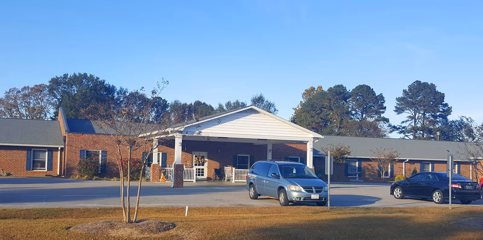Exterior view of a single-story brick building with a covered entrance supported by white columns. Two vehicles are parked in front of the building, and there are trees and a clear blue sky in the background.