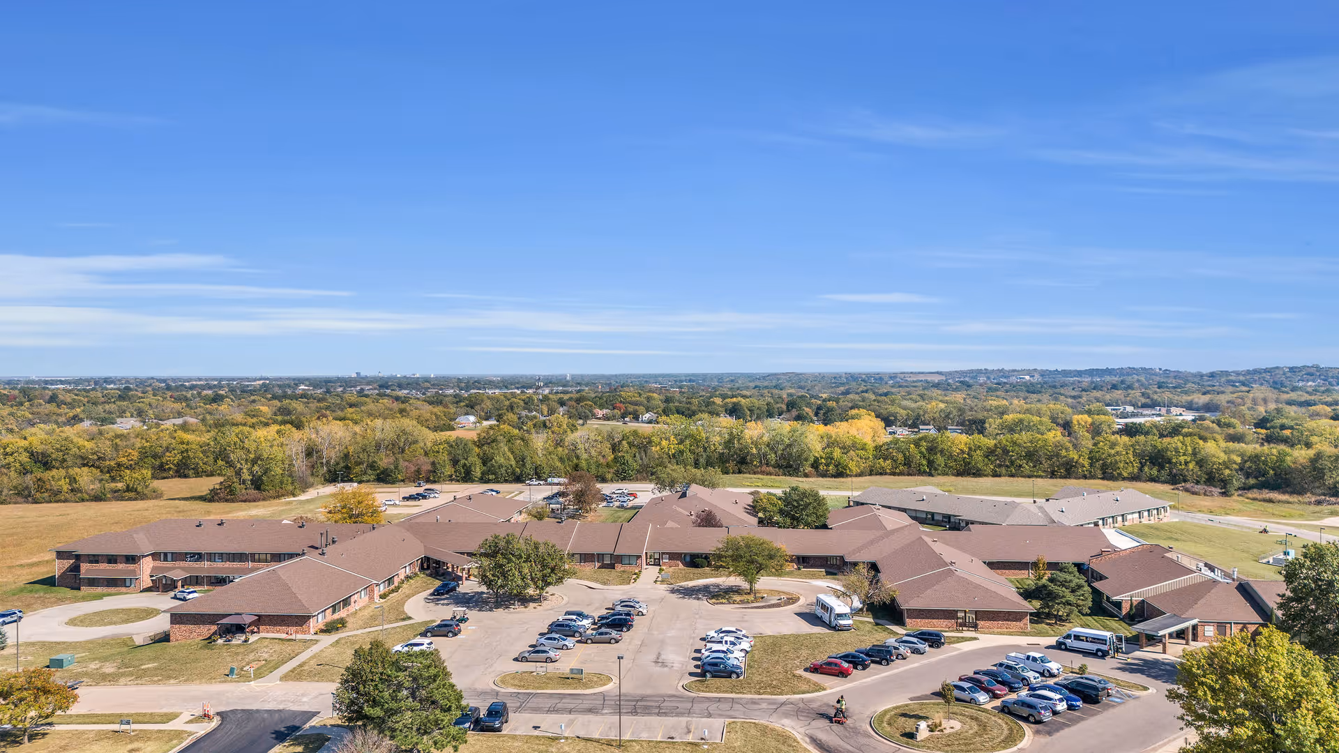 Aerial view of The Gardens at Aldersgate complex showing multiple low-rise buildings, parking lots, and surrounding trees and fields.