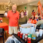 An elderly man and a middle-aged woman smiling and posing together in a decorated dining area with red tablecloths and festive decorations. In the foreground, there is a table with various beverages including soda cans, a bottle of wine, and a white ice bucket. Other people are seated and standing in the background.