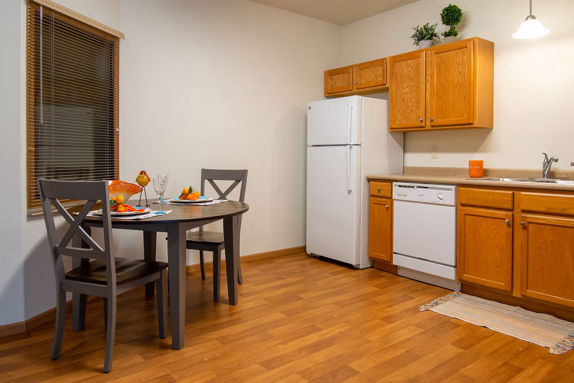 A small kitchen and dining area with wooden flooring and light-colored walls. The kitchen features wooden cabinets, a white refrigerator, a dishwasher, and a sink with a faucet. A small dining table with two chairs is set with plates, glasses, and decorative items near a window with wooden blinds.