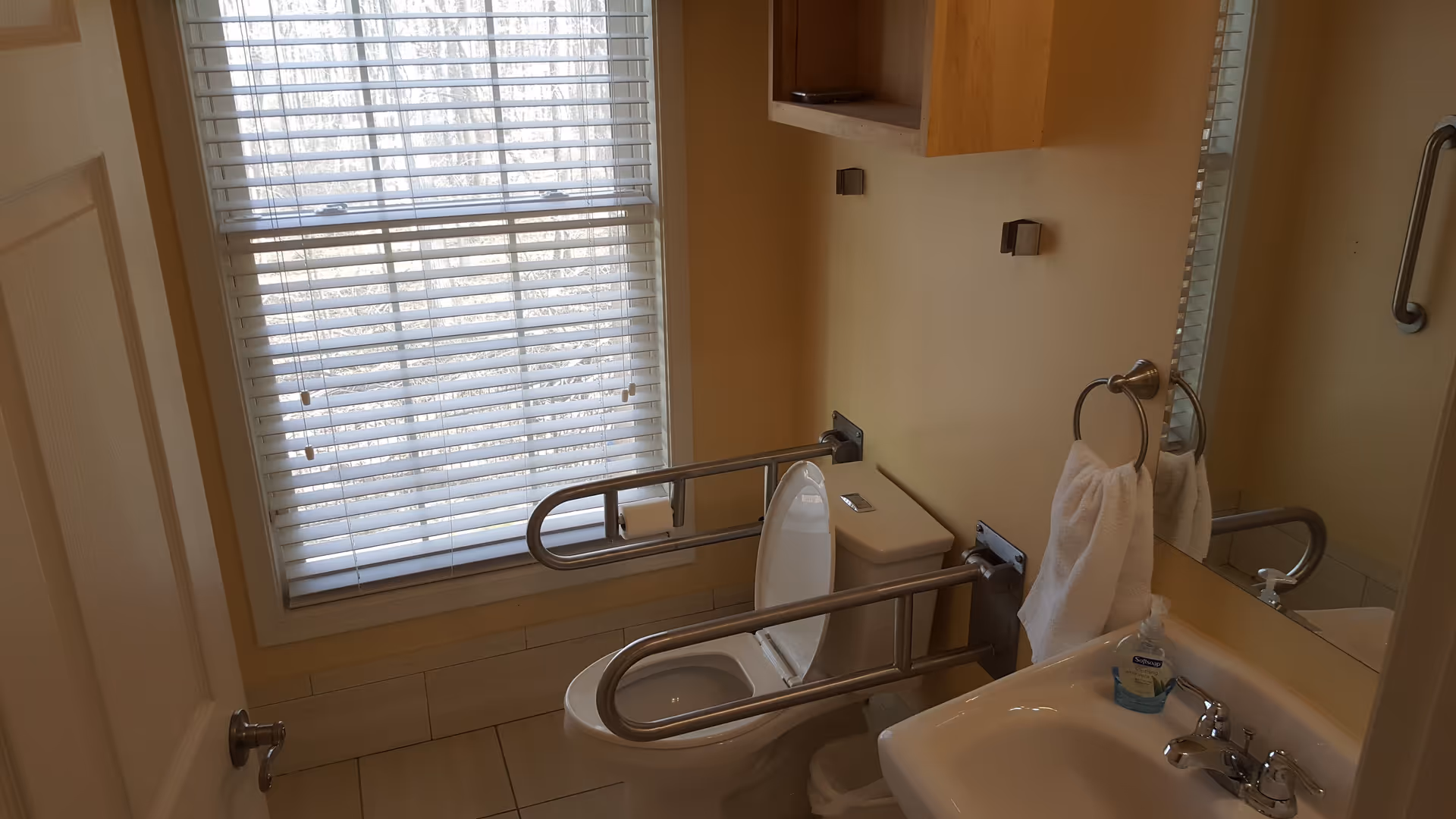 A small bathroom with a toilet equipped with metal safety rails on both sides, a white sink with a towel hanging on a ring holder, a bottle of hand soap on the sink, a large mirror above the sink, and a window with closed blinds letting in natural light.