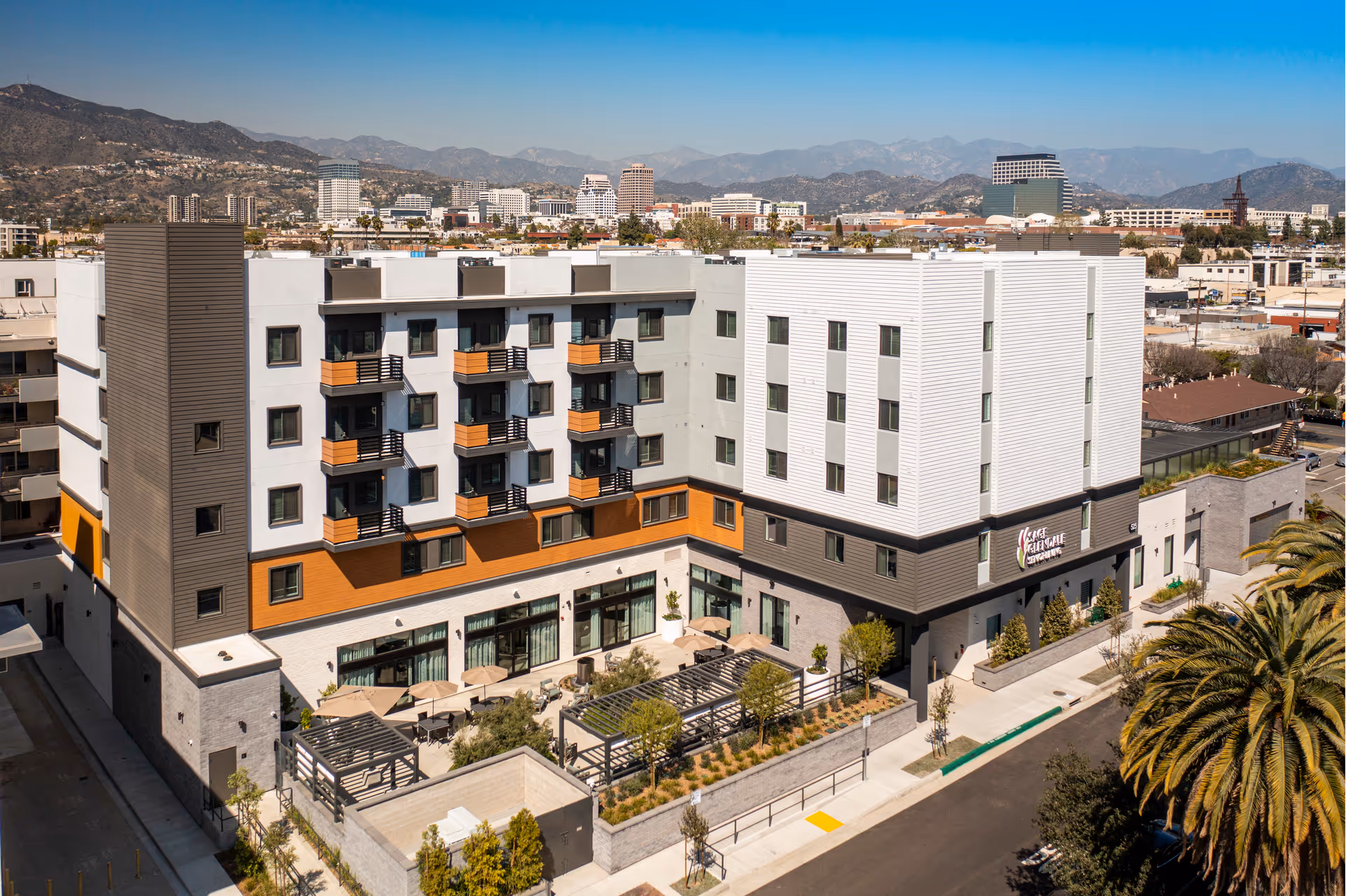 Aerial view of a modern multi-story senior living facility named Sage Glendale with outdoor patio areas featuring tables, chairs, umbrellas, and pergolas. The building has a mix of white, gray, and wood-tone exterior finishes. In the background, there is a cityscape with mountains under a clear blue sky.