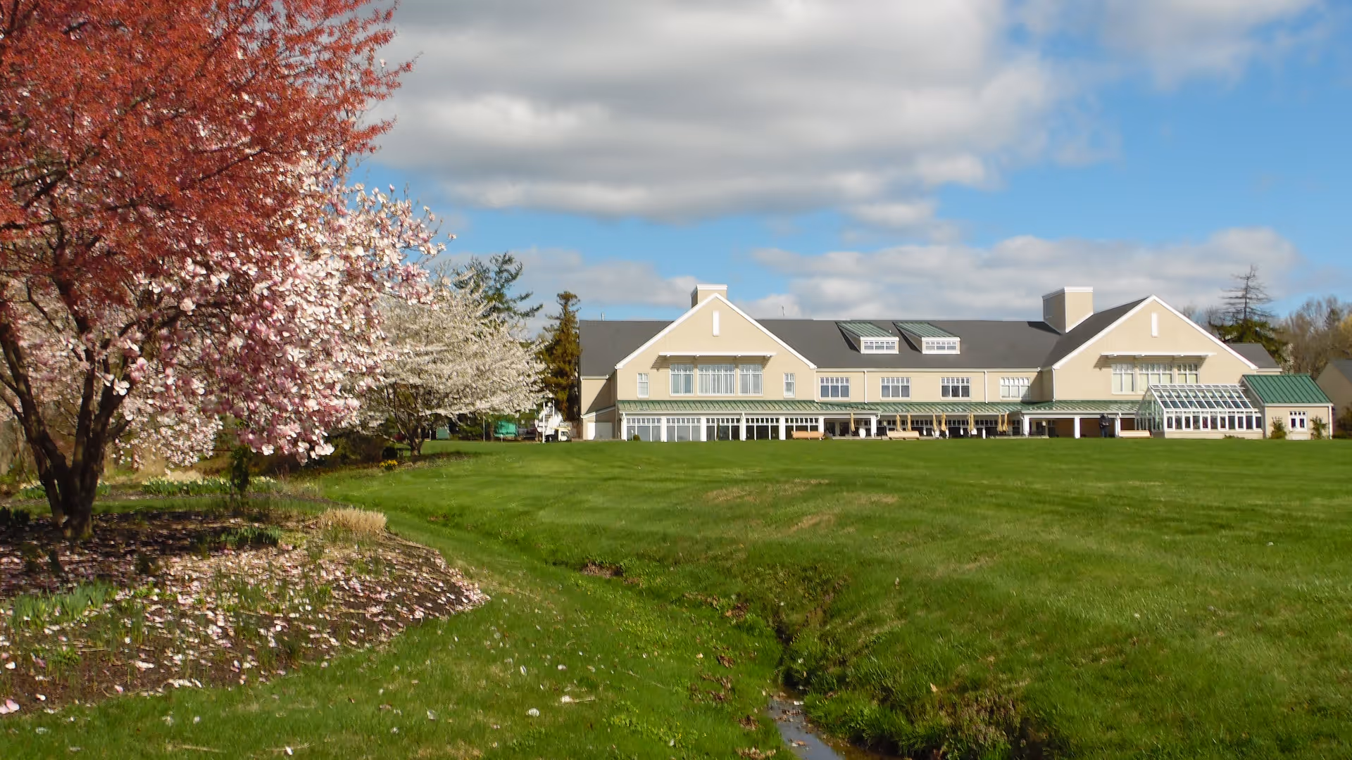 A large beige building with a dark roof and multiple windows, situated behind a wide green lawn with a small stream running through it. On the left side, there are blooming trees with pink and white flowers under a partly cloudy blue sky.