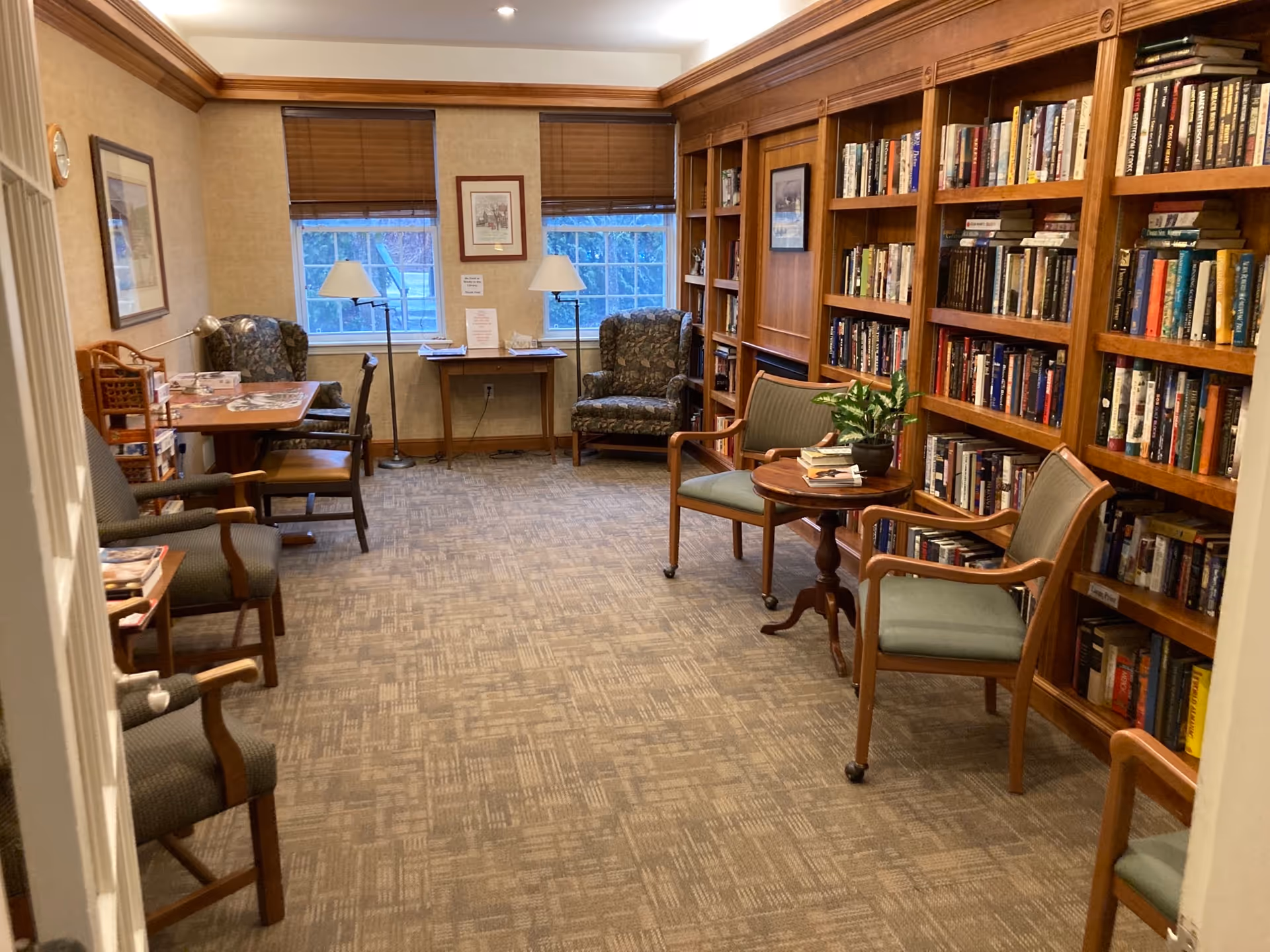 A cozy library room with carpeted floor, wooden bookshelves filled with books along the right wall, several chairs including armchairs and wooden chairs with cushions, a small round table with a plant and books, two windows with brown blinds, two floor lamps, and framed pictures on the walls.