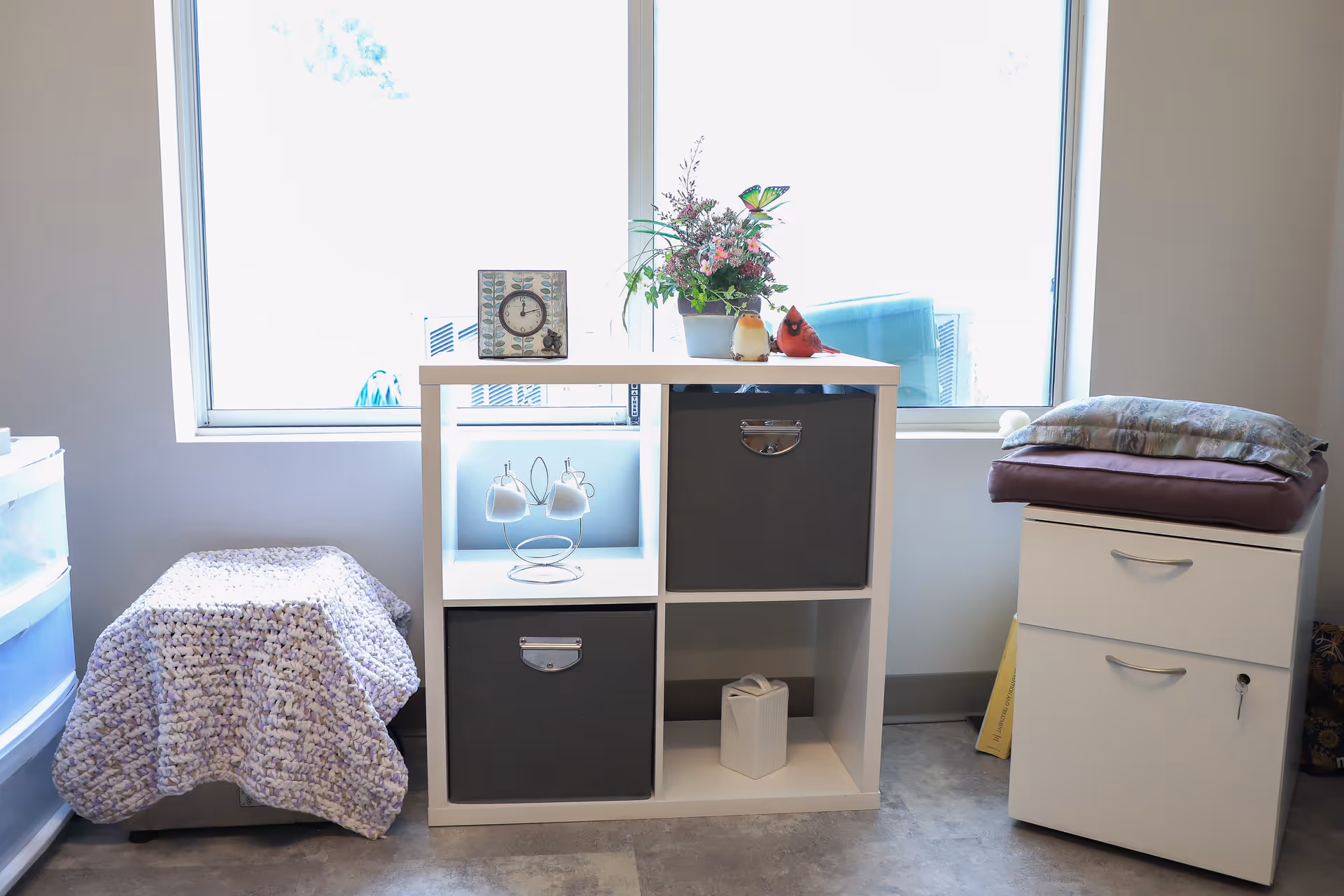 A white cubical shelf unit with two gray storage bins, a small clock, a flower arrangement, and two bird figurines on top, placed in front of a large window. To the left is a plastic drawer unit covered with a purple knitted blanket, and to the right is a white cabinet with two drawers, topped with folded blankets and a pillow.