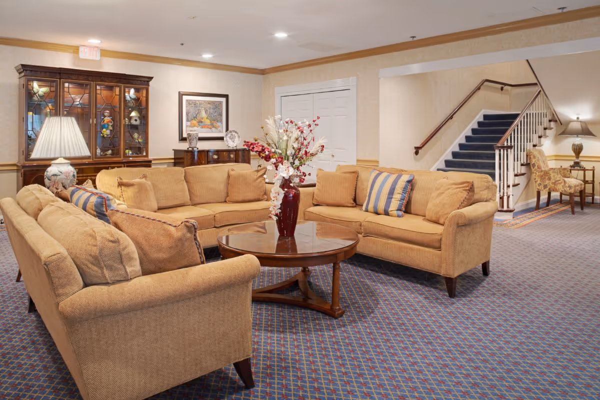 A cozy living room area with beige sofas arranged around a round wooden coffee table holding a vase with flowers. The room has patterned carpet flooring, a wooden display cabinet with glass doors, a framed painting on the wall, and a staircase with blue carpeting leading upstairs. There is also a side table with a lamp and an upholstered chair near the staircase.