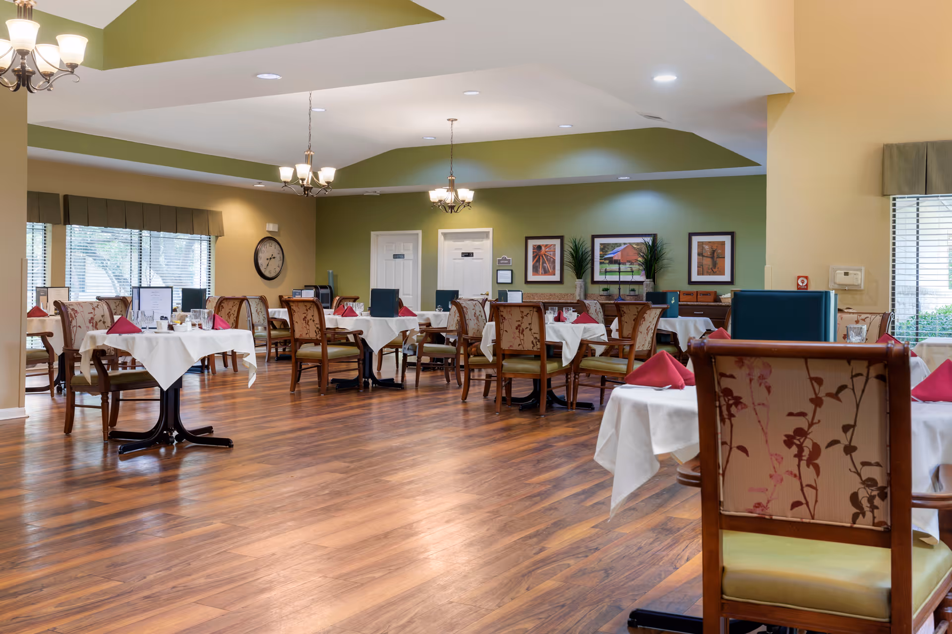 Spacious dining room with tables set with white tablecloths and red napkins, wooden chairs, chandeliers, and hardwood floors.