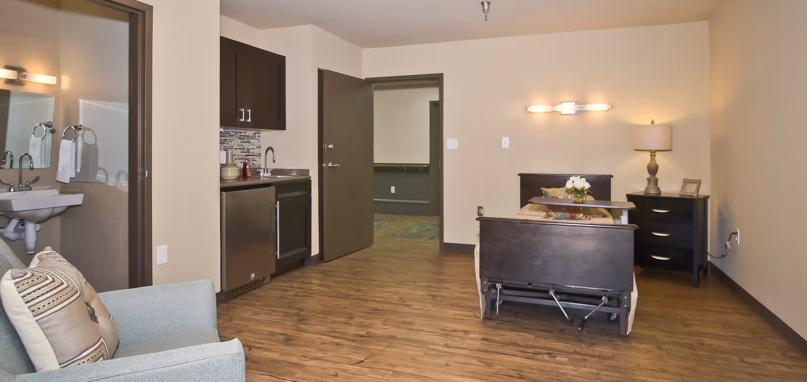 A senior living facility room featuring a hospital-style bed with a side table and lamp, a small kitchenette with a sink and cabinets, a comfortable armchair with pillows, and an open door leading to another room. The room has wood flooring and neutral-colored walls.