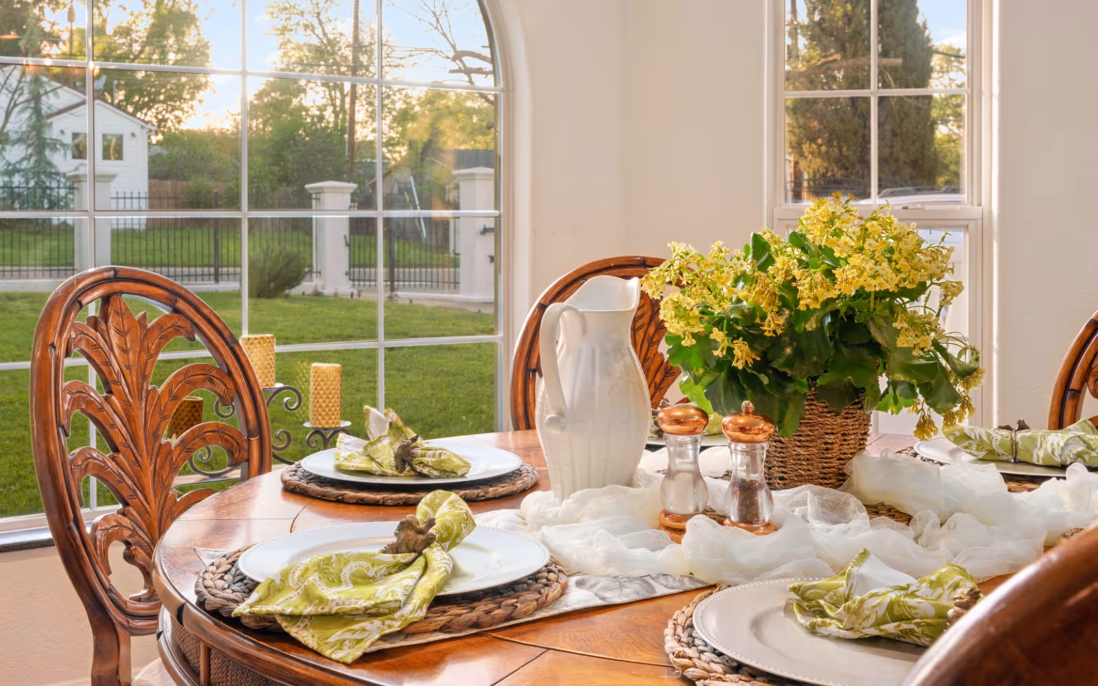 A wooden dining table set with white plates, green patterned napkins, a white pitcher, salt and pepper shakers, and a basket with yellow flowers. The table is near large windows showing a green lawn and trees outside.