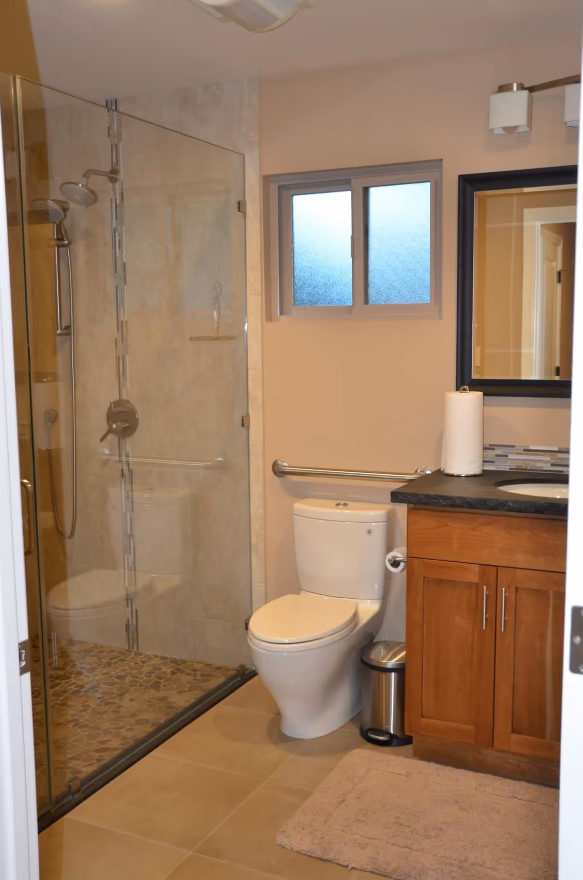 A clean and modern bathroom featuring a glass-enclosed shower with a pebble stone floor, a white toilet with a grab bar on the wall behind it, a wooden vanity with a black countertop, a sink, a mirror above the sink, a paper towel roll on the counter, a small trash can, and a beige bath mat on the tiled floor.