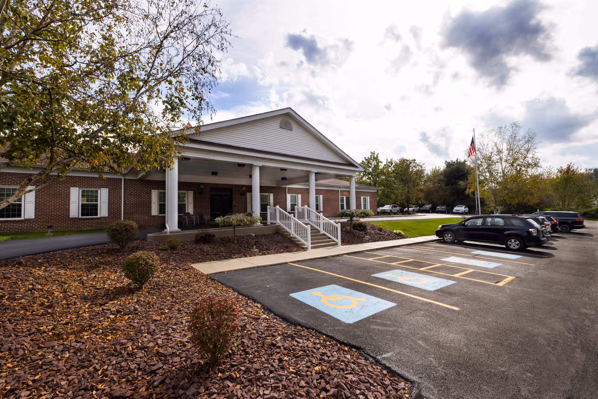 Exterior view of a single-story brick building with white columns and a covered entrance. There are several handicap parking spaces in the foreground, a few cars parked, and trees with autumn foliage surrounding the area under a partly cloudy sky.