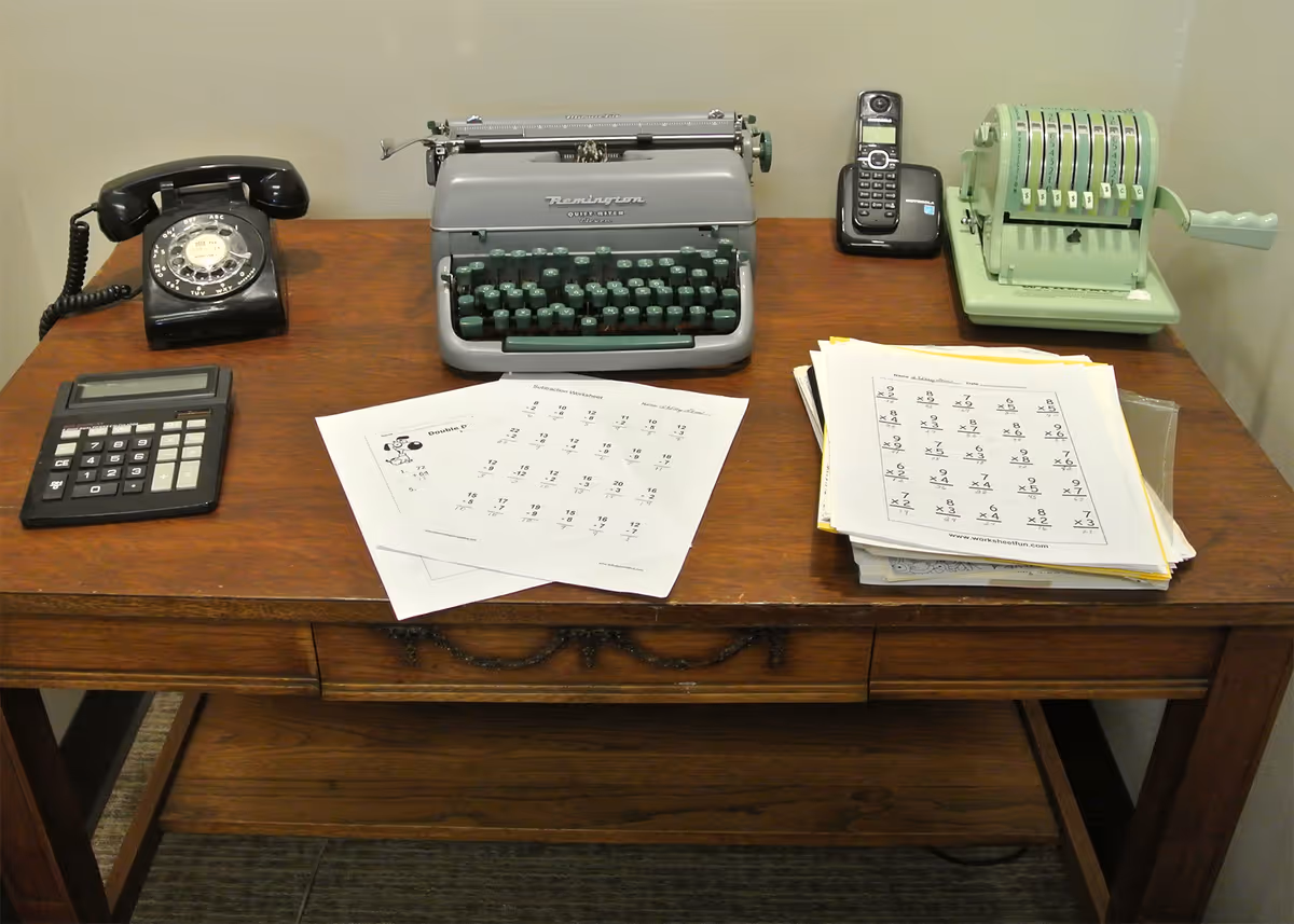 A wooden desk holding a vintage typewriter, rotary phone, calculator, adding machine, cordless phone and stacks of papers.