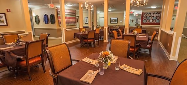 A warm and inviting dining room with several tables covered in brown tablecloths, each set with napkins, silverware, glasses, and small floral centerpieces. The room features wooden floors, beige walls with framed artwork and quilts, and soft lighting from chandeliers and ceiling fixtures.