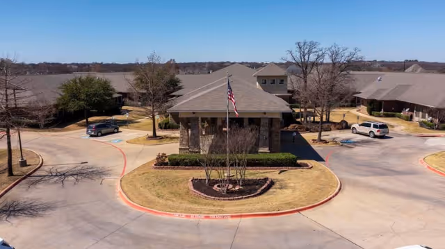 Aerial view of a healthcare facility entrance with a circular driveway, landscaped center island with an American flag, and surrounding buildings with pitched roofs. Several cars are parked near the buildings, and leafless trees are visible around the property under a clear blue sky.