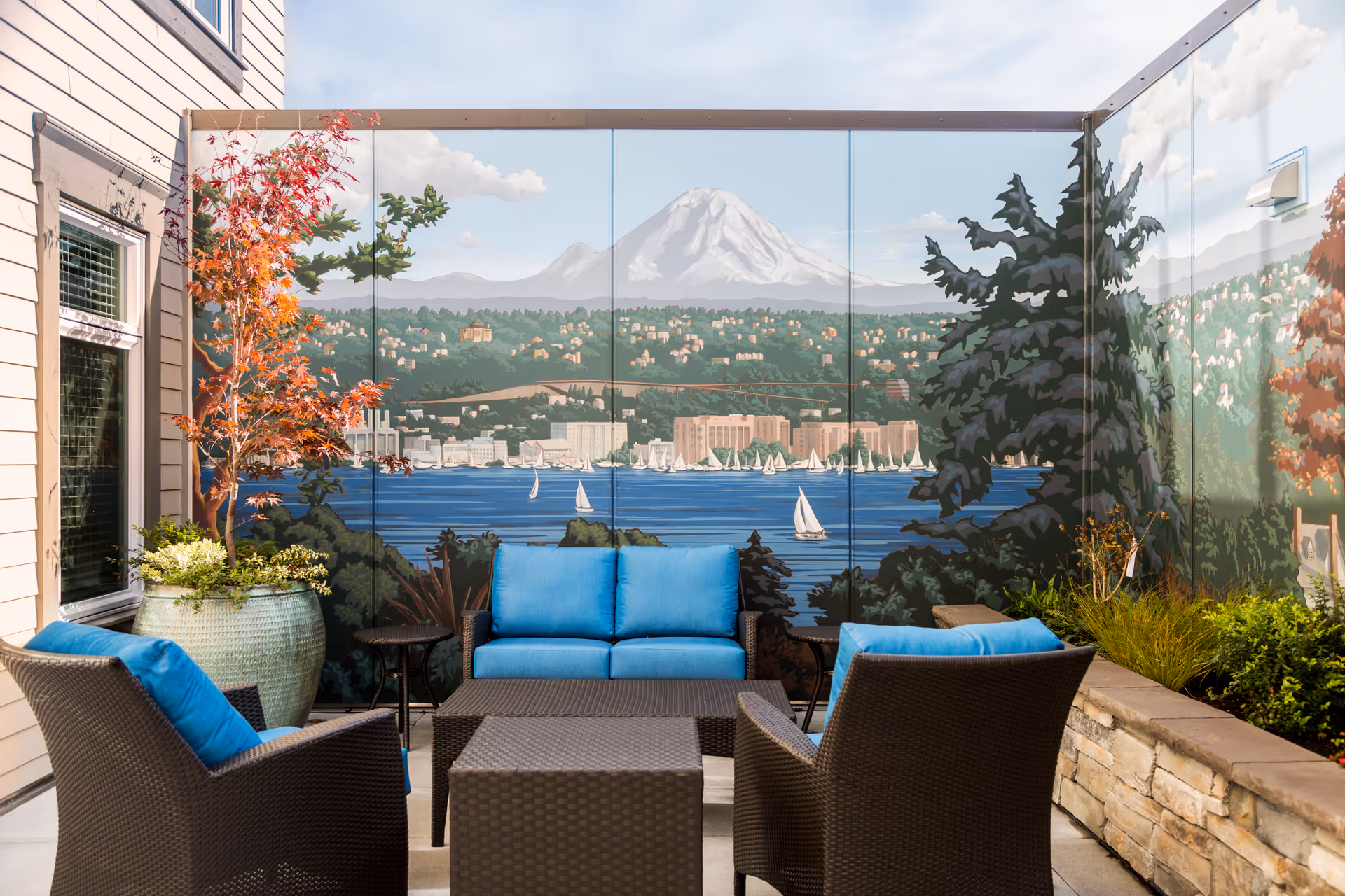 Outdoor patio seating area with wicker chairs and blue cushions in front of a large mural depicting a lake, sailboats and a mountain.