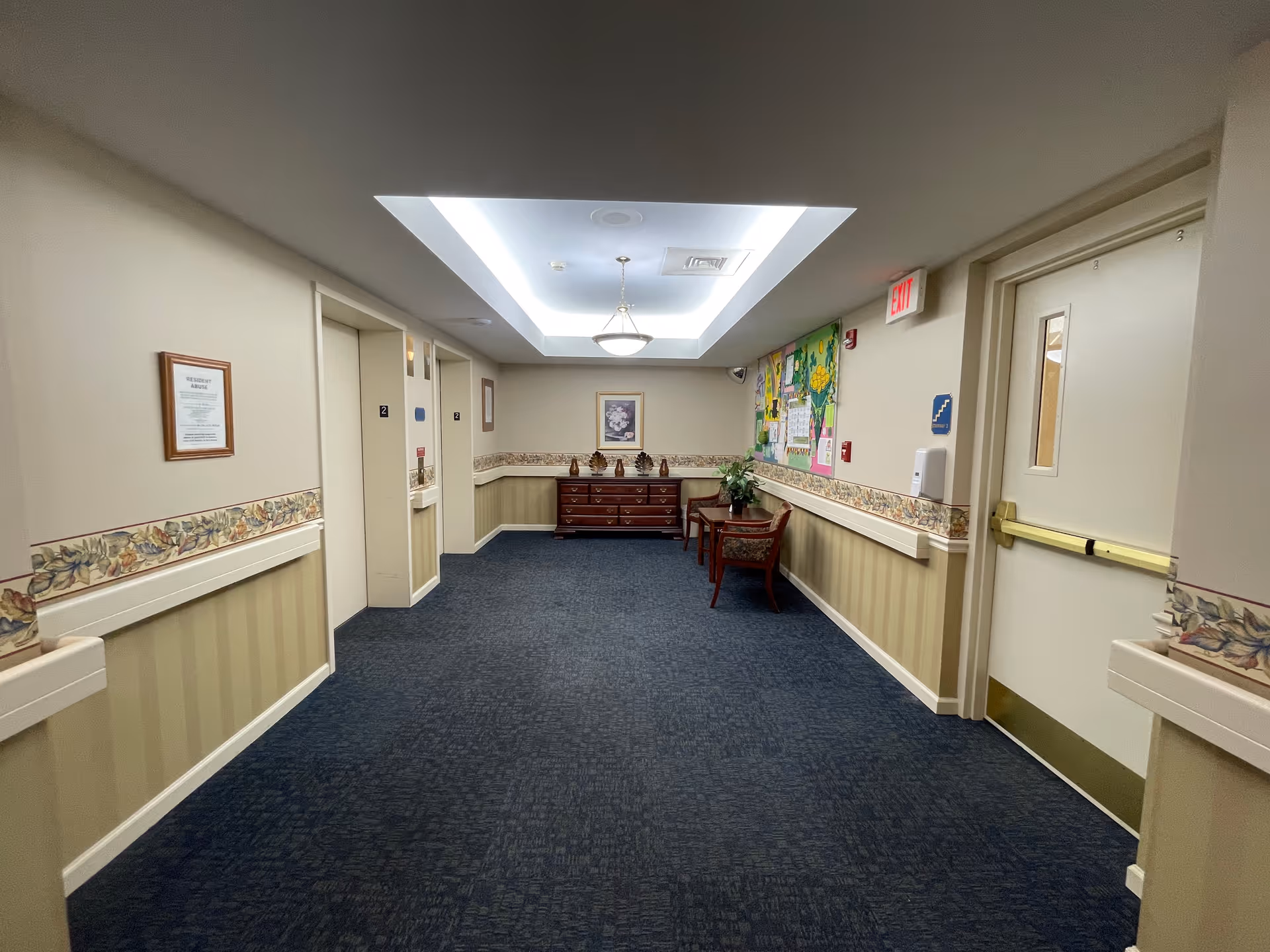 A well-lit hallway in a senior living facility with blue carpet and beige walls decorated with floral wallpaper borders. There are two elevators on the left side, a wooden dresser with decorative items and a framed picture on the far wall, and two chairs with a small table and plant on the right side. An exit sign is visible above a door on the right.