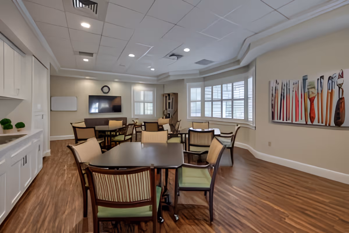 A well-lit dining room with several tables and chairs arranged on a wooden floor. The room features large windows with white blinds, a wall-mounted TV, a whiteboard, and a colorful abstract painting on the wall. The ceiling has recessed lighting and a decorative tray design.