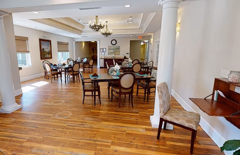 A bright dining room with wooden floors, several tables set with napkins and chairs, chandeliers hanging from the ceiling, and large windows letting in natural light. There are decorative columns and a small desk with a chair against the wall.