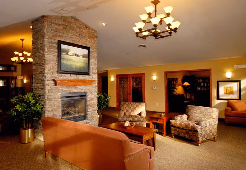 A cozy living room area in a senior living facility with a stone fireplace at the center. The room features a brown sofa, two patterned armchairs, a wooden coffee table, and side tables with lamps. Warm lighting comes from ceiling chandeliers and wall sconces, and there are framed pictures on the walls. Potted plants add greenery to the space.