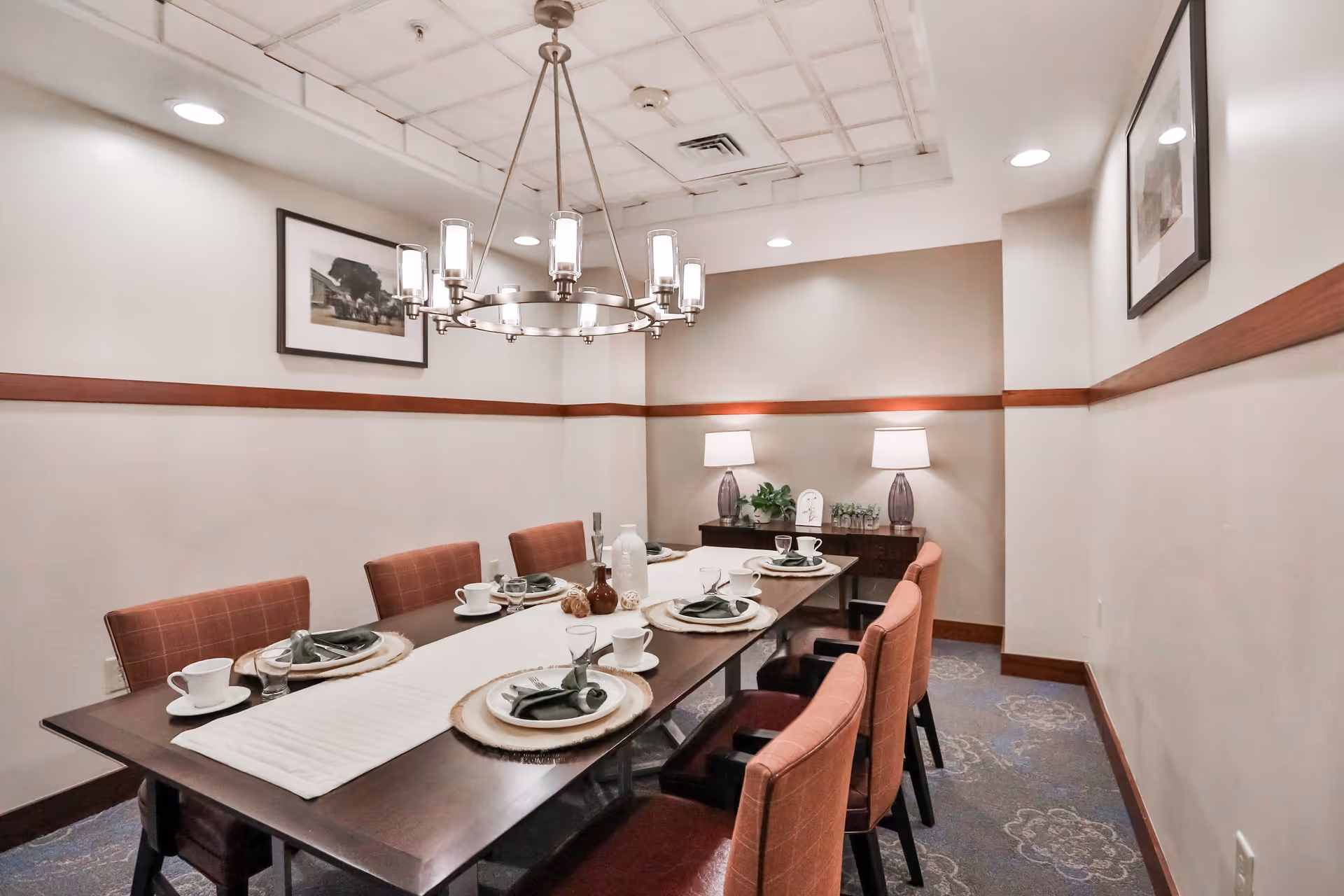 A dining room with a long rectangular wooden table set for six people. The table has white cups, plates with green napkins, and a white table runner. There are six upholstered chairs around the table. The room has beige walls with a wooden trim, two framed pictures on the walls, a chandelier hanging from the ceiling, and a sideboard with two lamps and decorative items.