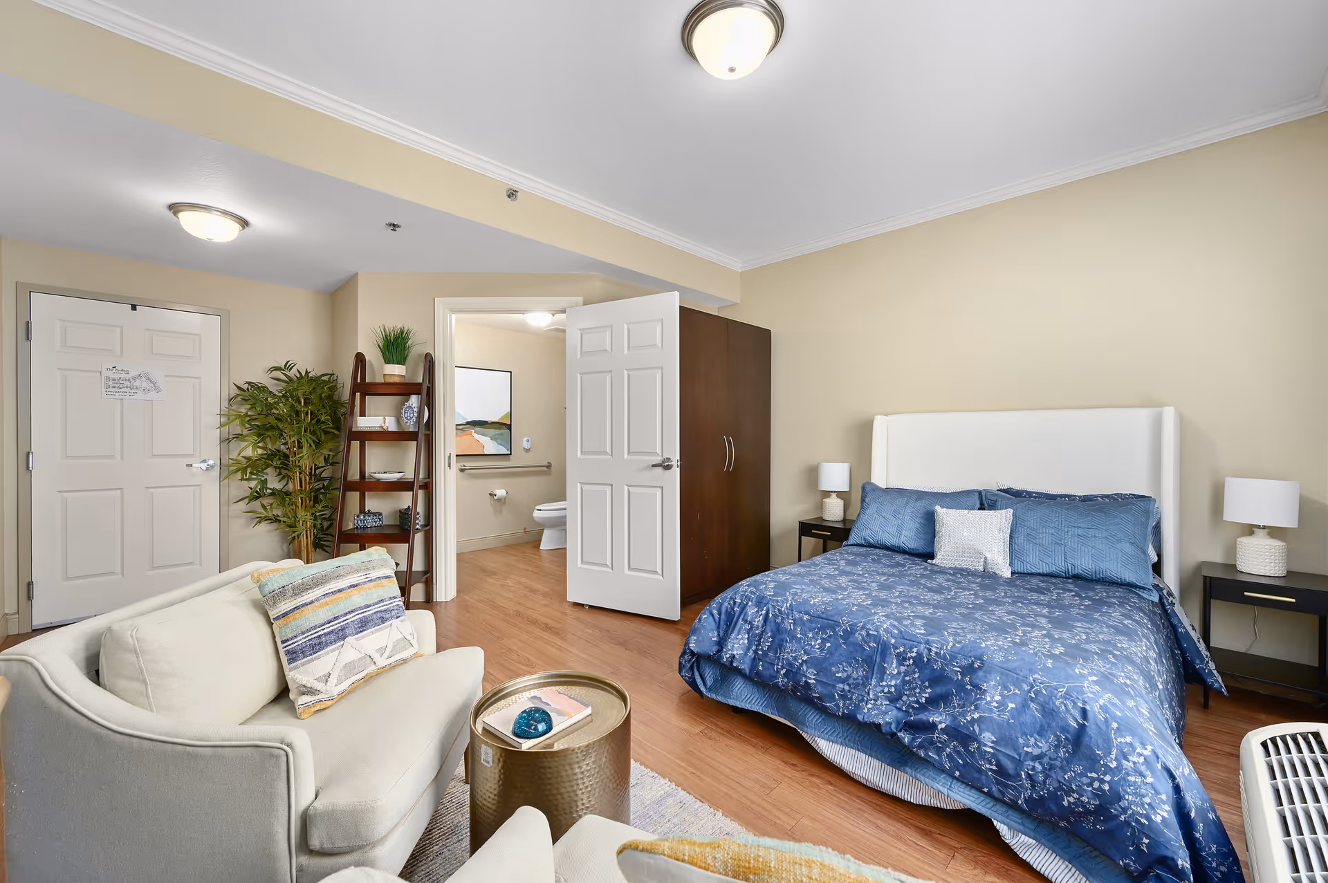 Studio apartment bedroom with a blue-dressed bed, a seating area, shelving, and an open door to a bathroom.