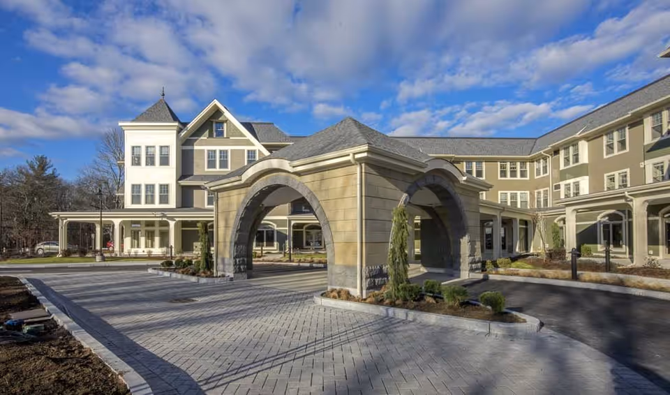 Exterior view of a large senior living facility building with a covered driveway entrance featuring stone arches, surrounded by landscaped grounds under a partly cloudy sky.