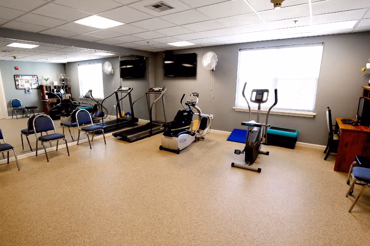 A fitness room with exercise equipment including two treadmills, a recumbent bike, and an elliptical machine. The room has a large mirror on one wall, two wall-mounted fans, two flat-screen TVs, several chairs arranged along the walls, and a desk with a computer in the corner. The room is well-lit with natural light coming through a window with blinds.