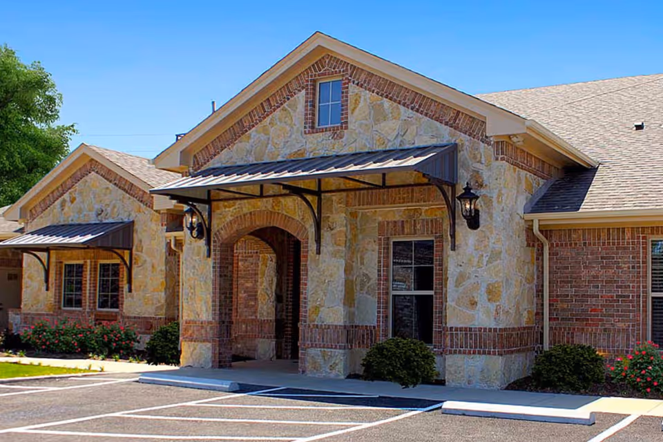 Exterior view of a single-story building with stone and brick facade, featuring two entrances with metal awnings and outdoor wall lanterns. There are small bushes and flowers planted along the building, and a parking lot with marked spaces in front.