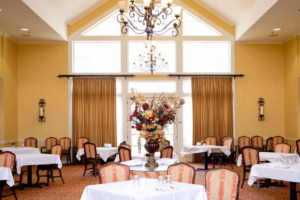 Formal dining room with tables covered in white linens, upholstered chairs, a large floral centerpiece, and chandeliers.