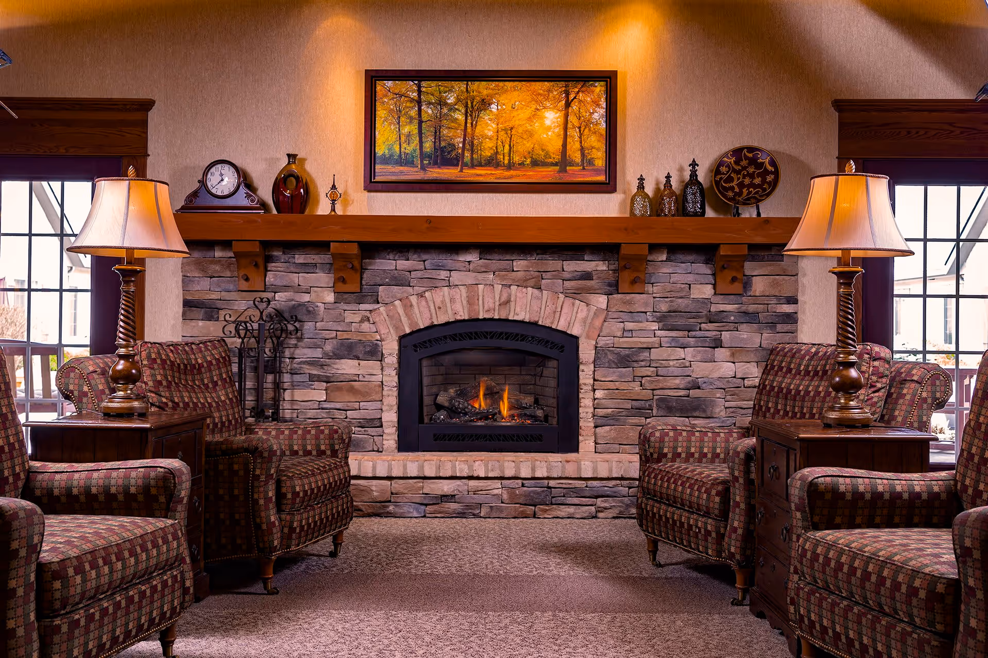 A cozy living room area with a stone fireplace in the center, a fire burning inside. Above the fireplace is a framed picture of a forest with autumn foliage. On either side of the fireplace are two windows with wooden frames. Four patterned armchairs are arranged around two wooden side tables, each with a lamp. Decorative items including a clock, vases, and a plate are placed on the wooden mantel above the fireplace.